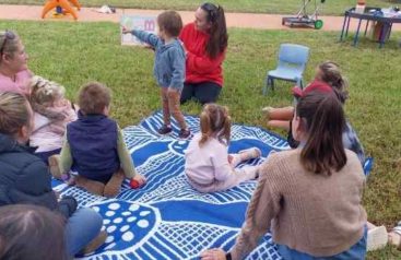 A group of children and adults in a park gathered around a person reading a book