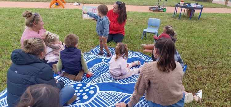 A group of children and adults in a park gathered around a person reading a book